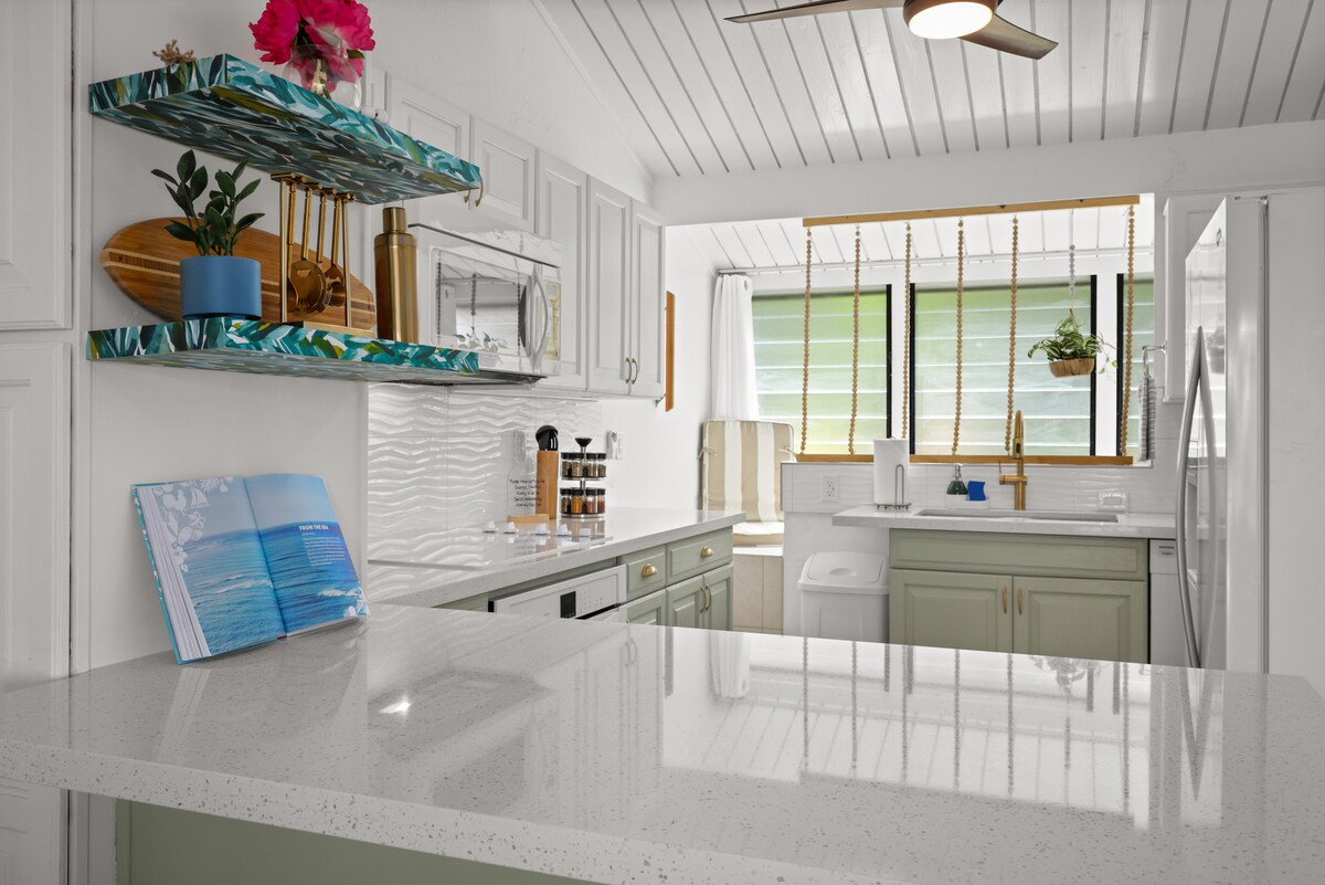 A modern kitchen is highlighted by a clean design featuring white cabinetry and a green accent on the lower cabinets. A large island with a polished countertop provides ample workspace. Natural light streams through the window, illuminating the well-equipped kitchen amenities.