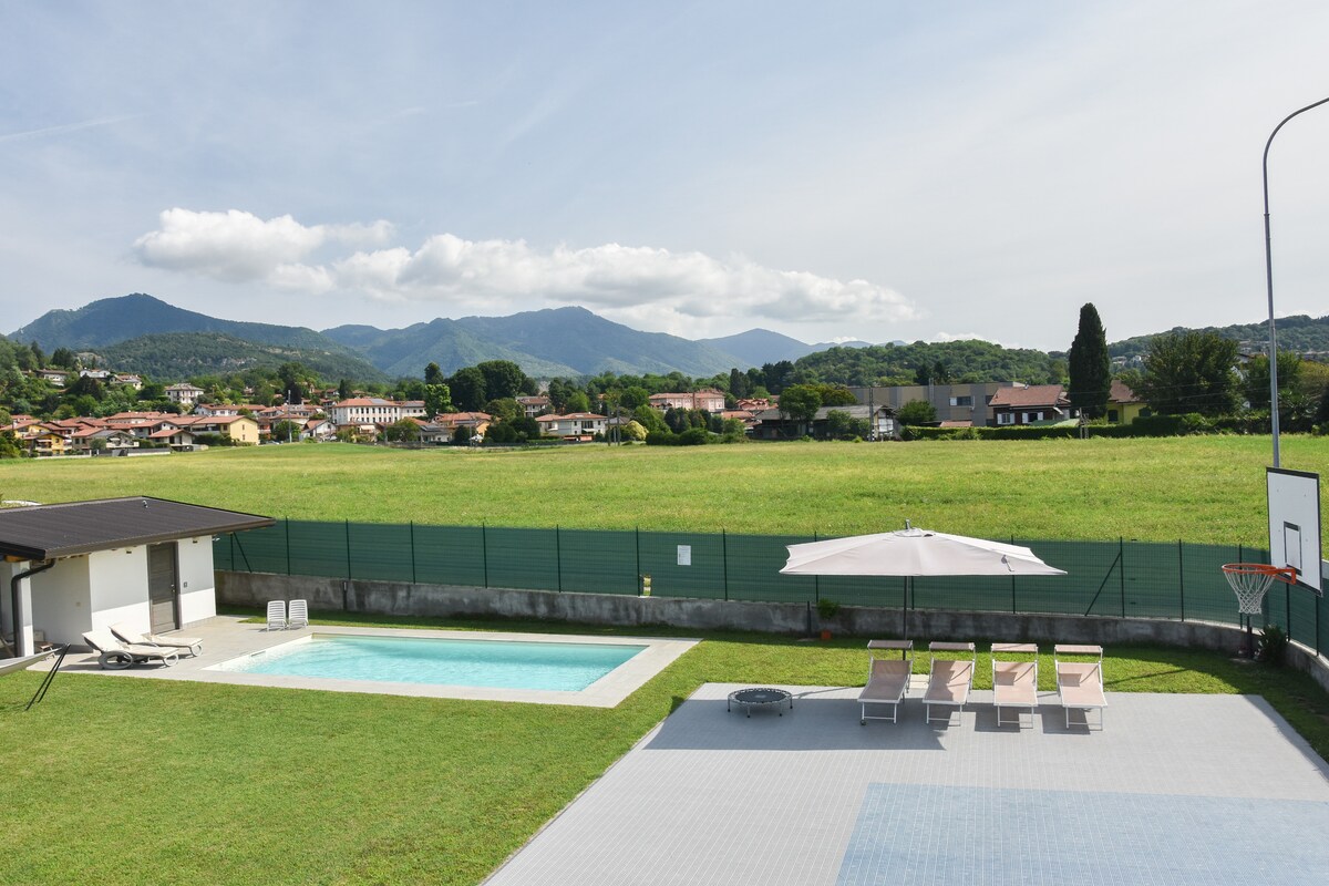 A serene outdoor area features a swimming pool surrounded by a grassy lawn and sun loungers. An umbrella provides shade near the pool. In the background, rolling hills and distant mountains create a natural setting, while a basketball hoop is visible to the right.
