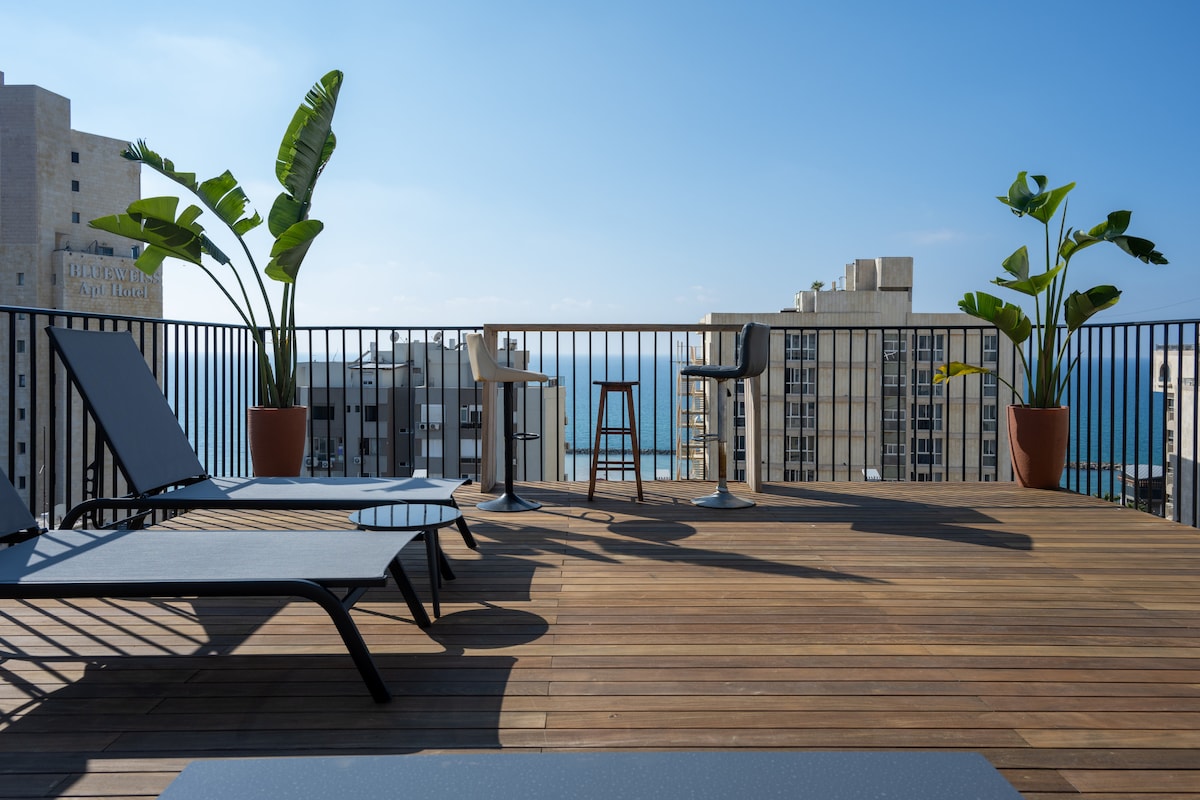 A spacious terrace features modern sunbeds and chic seating arrangements. Lush potted plants add a touch of greenery, while panoramic views of the sea and city create a serene backdrop. The wooden deck enhances the outdoor experience, inviting relaxation in the sun.