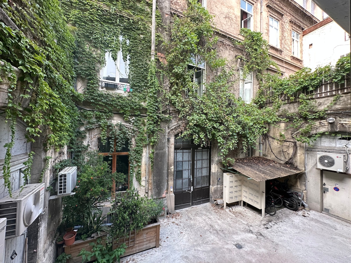 An inner courtyard is featured, surrounded by greenery and ivy-covered walls. Windows with plants in front provide a serene view. A covered area at the side holds bicycles, and air conditioning units are visible on the building's exterior, complementing the tranquil setting.