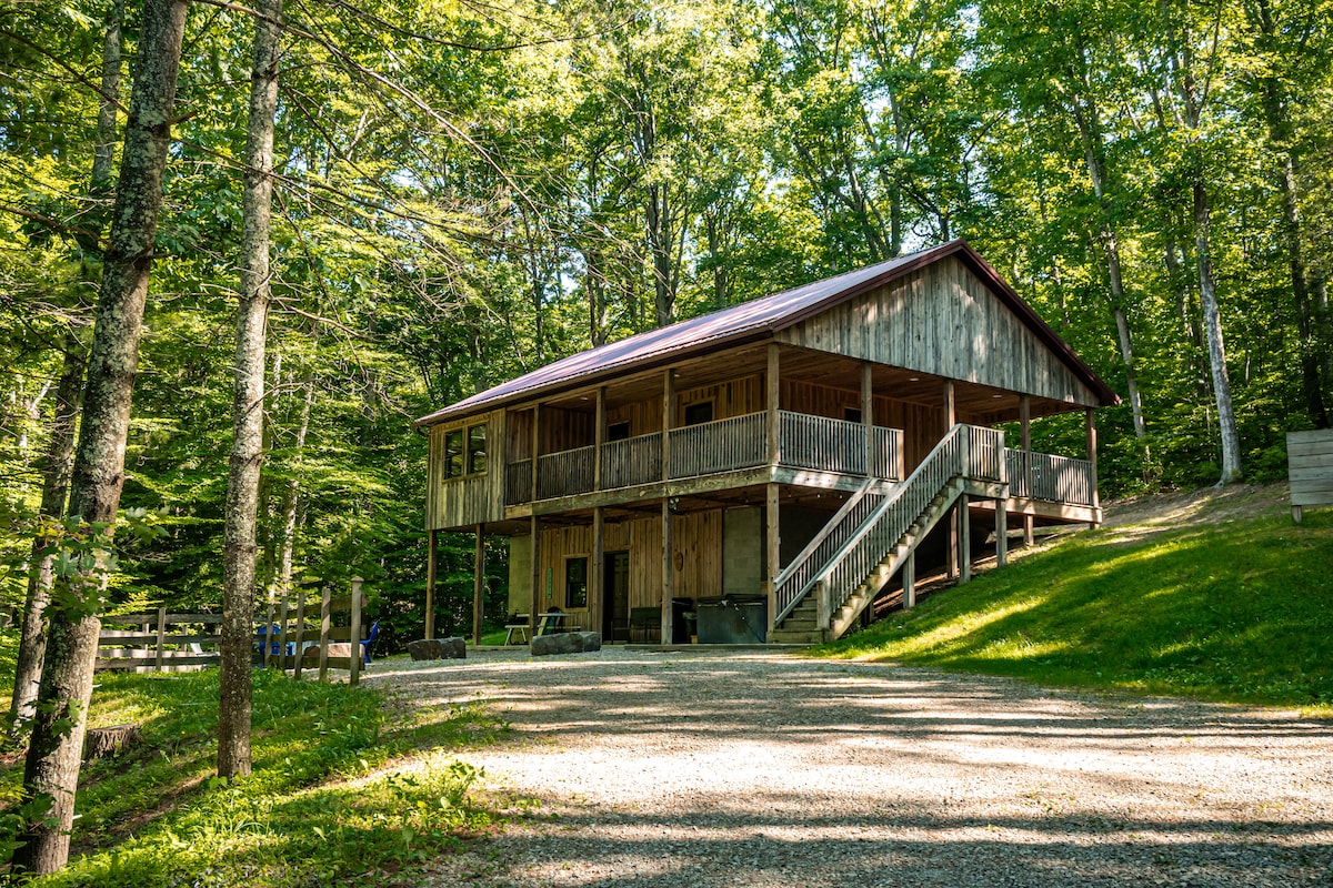 A secluded cabin is nestled among trees, featuring a wooden exterior and a covered wrap-around deck. The entrance is accessed by a set of stairs, with a gravel pathway leading up from the driveway, surrounded by lush greenery.