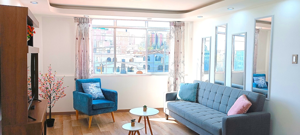 A well-lit living area is presented, featuring large windows that provide a view of the surrounding cityscape. A sofa complements two blue armchairs, with a small round table centrally positioned. Mirrors adorn the walls, reflecting natural light and enhancing the spacious feel of the room.