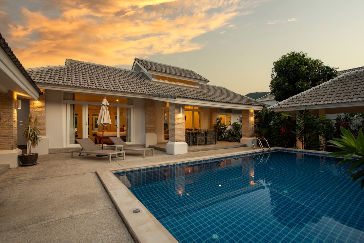 An outdoor area features a large swimming pool reflecting the warm colors of the sunset. Sun loungers are arranged beside the pool, while a dining table can be seen through wide glass doors leading into the villa. Lush greenery surrounds the space.