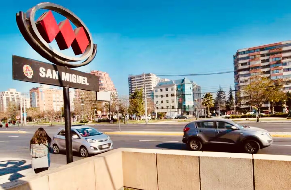 The image captures the San Miguel Metro station's sign, prominently displayed against a clear blue sky. A pedestrian strolls along the sidewalk while vehicles are seen moving on the road. Buildings with varying architectural styles can be observed in the background.