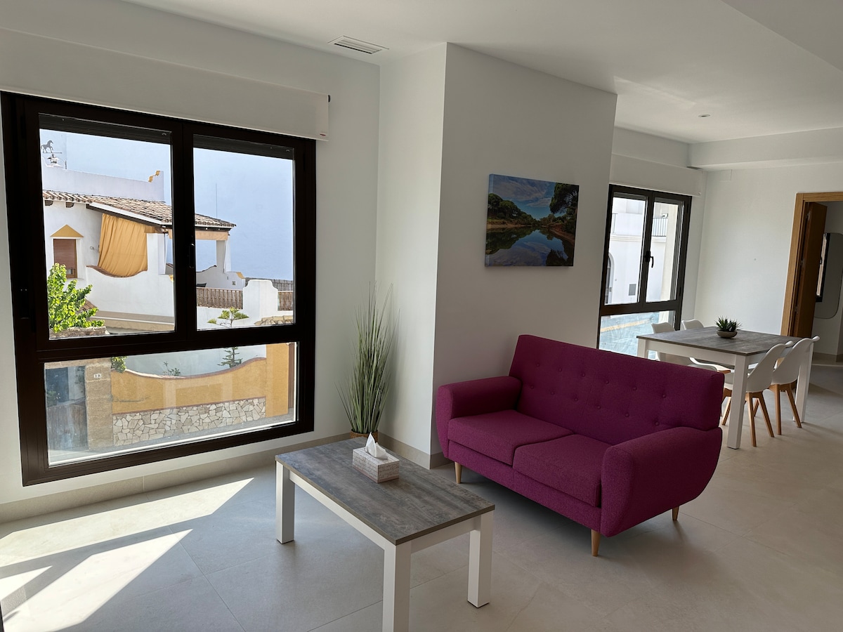 A bright living area features a pink sofa alongside a light wooden coffee table. Large windows provide natural light and views of the outdoors. A neutral-toned dining table is visible in the background, enhancing the spacious layout of the room.