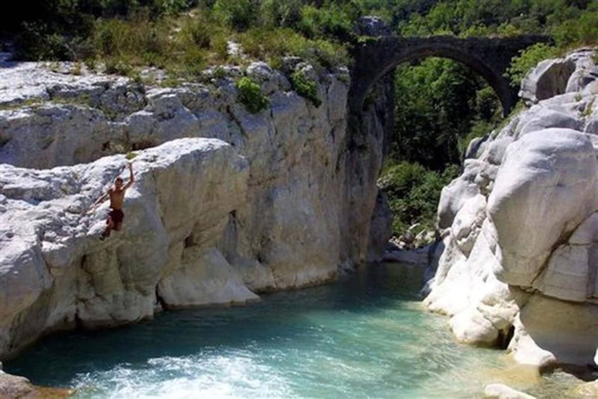 A clear, tranquil river is visible, with smooth, light-colored rocks lining its banks. A person can be seen preparing to jump into the water, which reflects the surrounding greenery. In the background, a stone arch bridge spans over the rocky terrain, surrounded by lush vegetation.