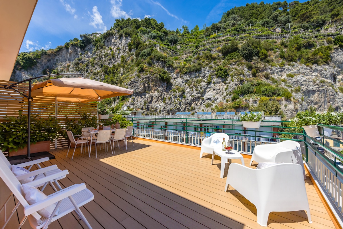 An expansive terrace offers multiple loungers and a dining area shaded by an umbrella. Lush greenery borders the space, with views of the surrounding cliffs and trees in the background. The wooden deck enhances the outdoor experience, inviting relaxation and connection with nature.