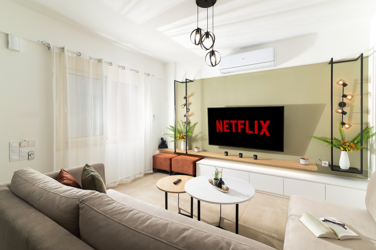 A bright living room features a large flat-screen TV displaying the Netflix logo, framed by decorative lighting and greenery. A neutral-toned sofa is accompanied by a round coffee table, while soft natural light filters in through sheer curtains, creating a comfortable and inviting space.