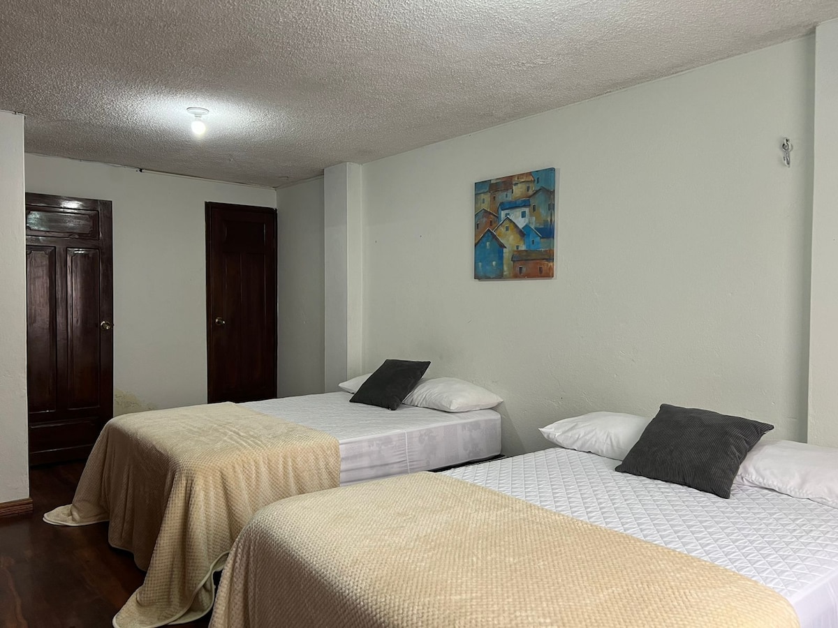 A spacious room features two neatly made beds covered with light blankets, complemented by decorative pillows. A colorful wall art piece depicting a landscape is displayed above the beds. Wooden doors are visible along the wall, adding warmth to the space.