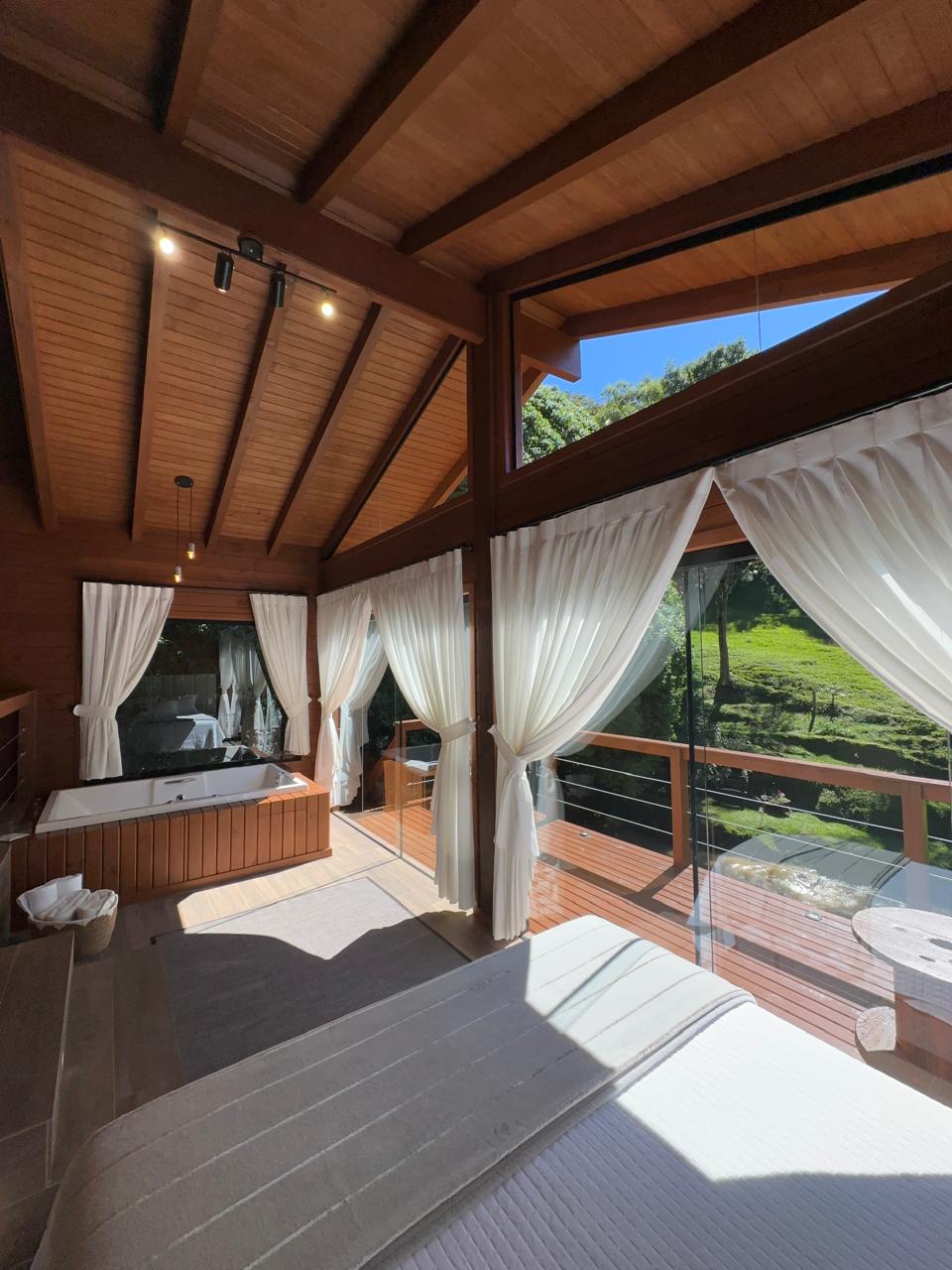 A spacious room is featured with wooden beams and large windows allowing natural light to flood in. White curtains frame the windows, creating a soft contrast with the wooden interior. A small outdoor area with greenery is visible through the glass doors.