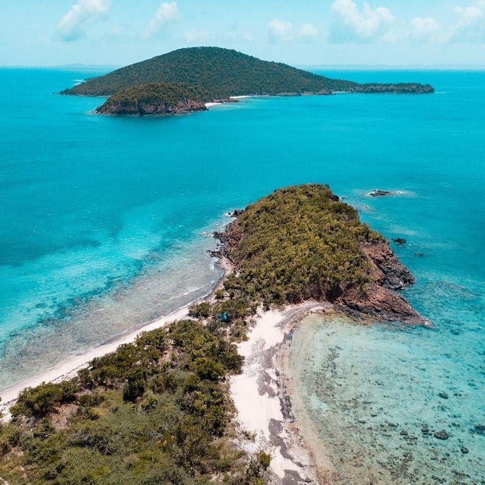An aerial view showcases a serene coastal landscape, featuring lush green hills on small islands surrounded by vibrant blue waters. Sandy shorelines curve gently, with sections of shallow water reflecting light. The scene presents a tranquil and inviting natural environment.