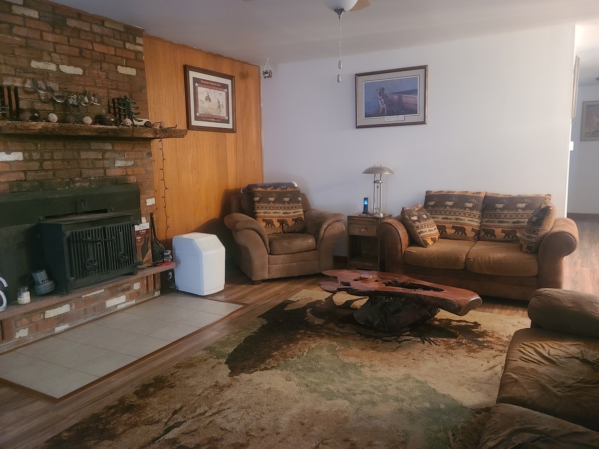 The living room features comfortable seating arrangements with two large couches and a loveseat, complemented by a rustic wooden coffee table. A brick fireplace is situated against the wall, and artwork adorns the space, creating a cozy gathering area.