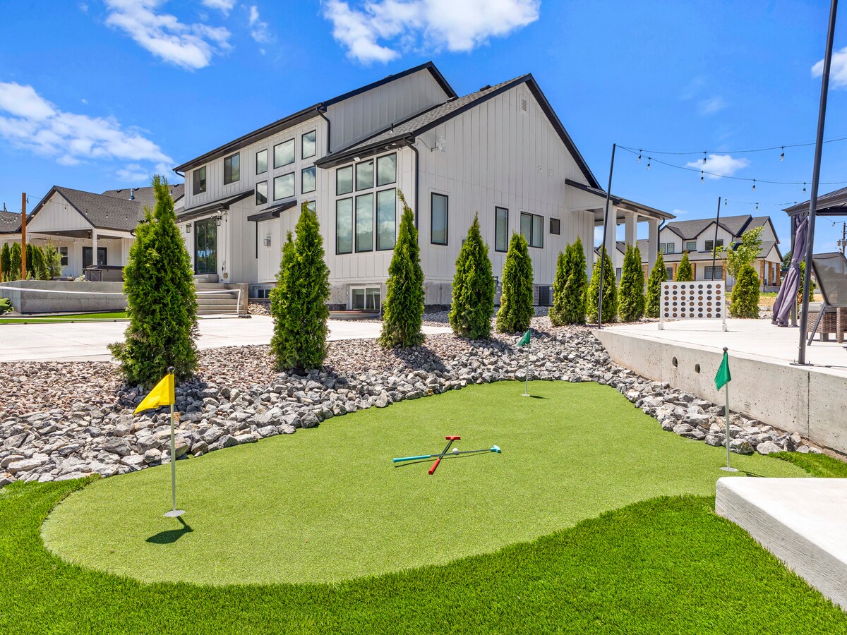 A custom putting green is set against a backdrop of a modern farmhouse, surrounded by neatly trimmed hedges. The vibrant green turf is accented with colorful flags and golf clubs positioned on the green, inviting outdoor leisure within the spacious yard.