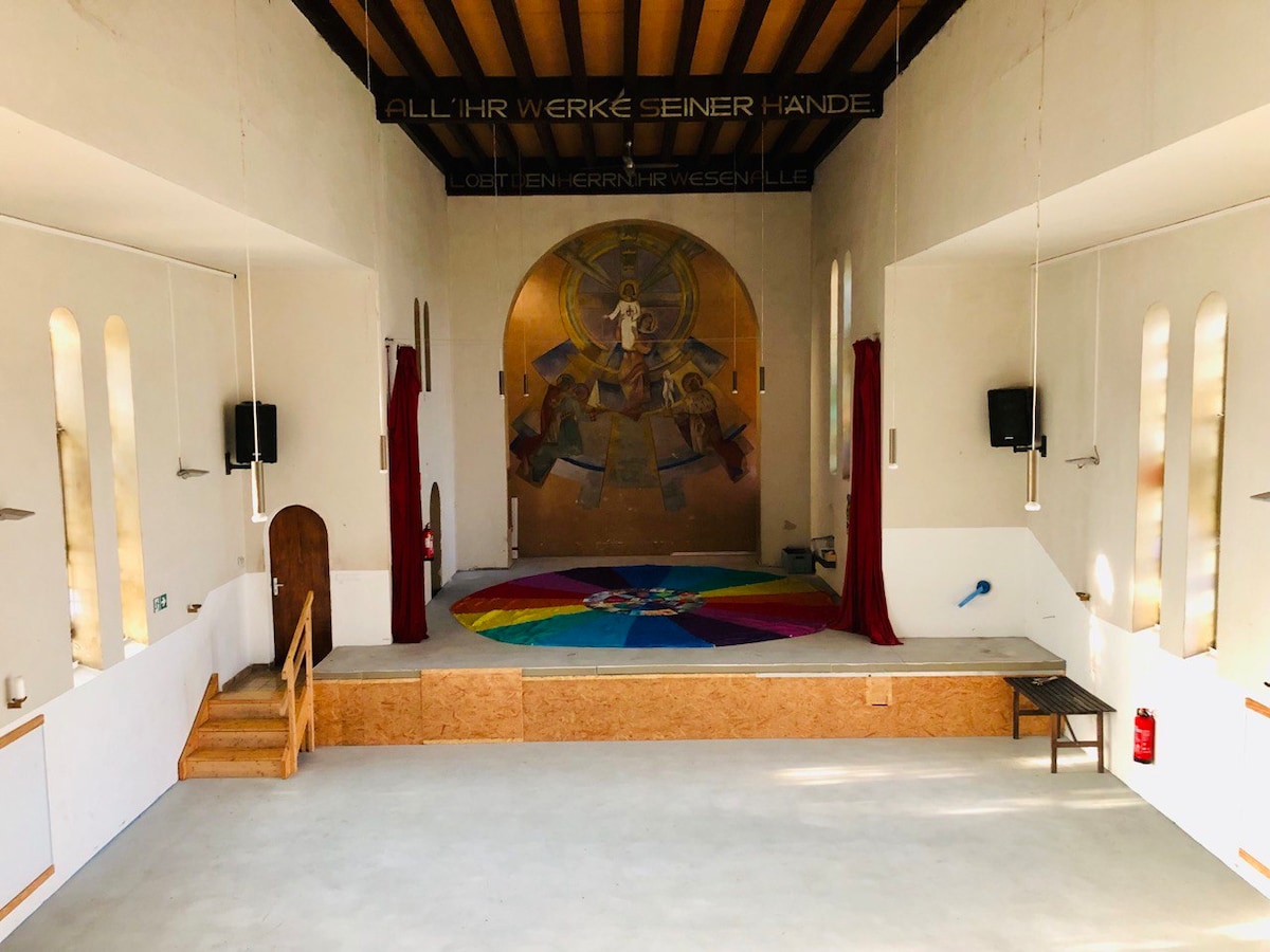 The former church interior features a spacious hall with high ceilings and large windows allowing natural light to fill the space. A colored circle design is centered on the floor, with wooden steps leading to the raised stage area. Red curtains and sound equipment are visible on the walls.