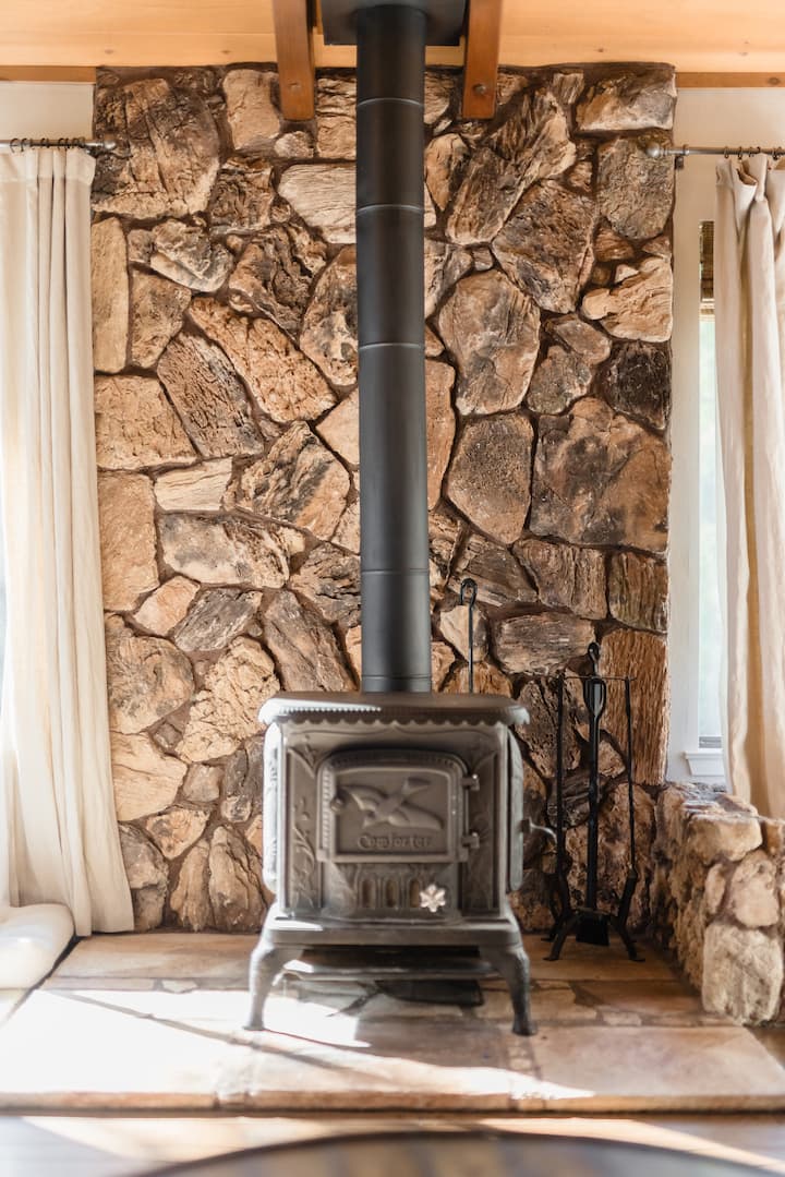 A black wood-burning stove set against a rustic stone wall. The stove has a pipe extending upwards and is flanked by beige curtains.