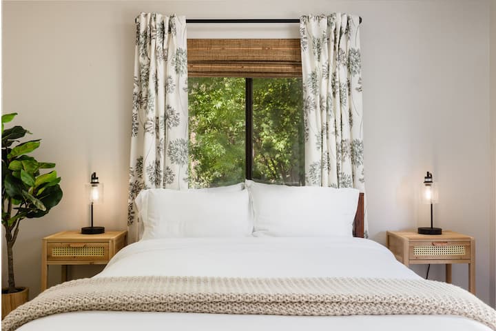 A serene bedroom with a white bed, wooden nightstands, lamps, and a large window with bamboo shades and patterned curtains.