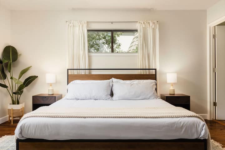 Master bedroom featuring king bed and two bedside tables with lamps. A small window with curtains is above the bed, and a potted plant is in the corner.
