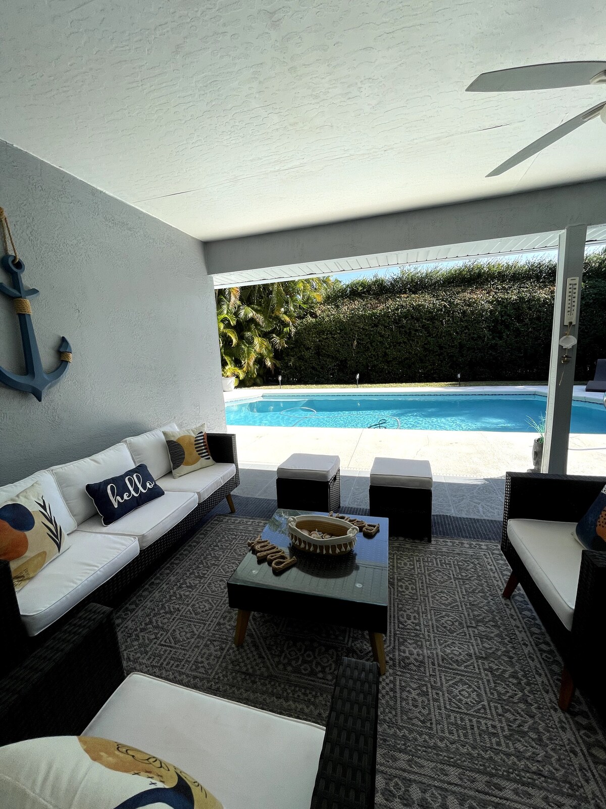 A shaded outdoor lounge area offers comfortable seating with neutral and blue cushions. A small coffee table is centered among the seating. The view beyond shows a clear pool surrounded by greenery, creating a tranquil setting.