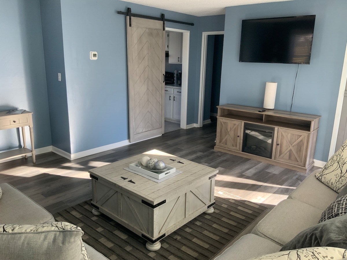 A light-filled living area features a spacious sofa and neutral-toned accents. A large flat-screen TV is mounted on the wall, with a wooden console below. A textured area rug lies beneath a coffee table adorned with decorative items, complemented by soft blue walls.