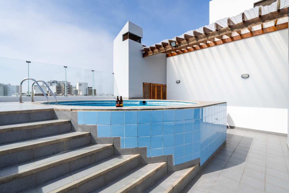 A modern rooftop pool area features a circular pool surrounded by light blue tiles and a sun-drenched terrace. Steps lead down to the pool, where partial views of the city skyline can be seen in the background under a clear blue sky.