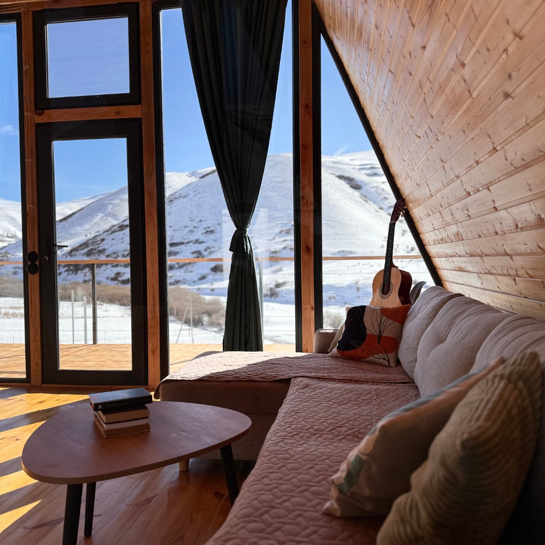 A spacious living area features a comfortable couch adorned with decorative pillows. A coffee table holds a stack of books. Large windows provide a stunning view of the snowy mountains outside, while light filters through dark curtains, enhancing the cozy ambiance.