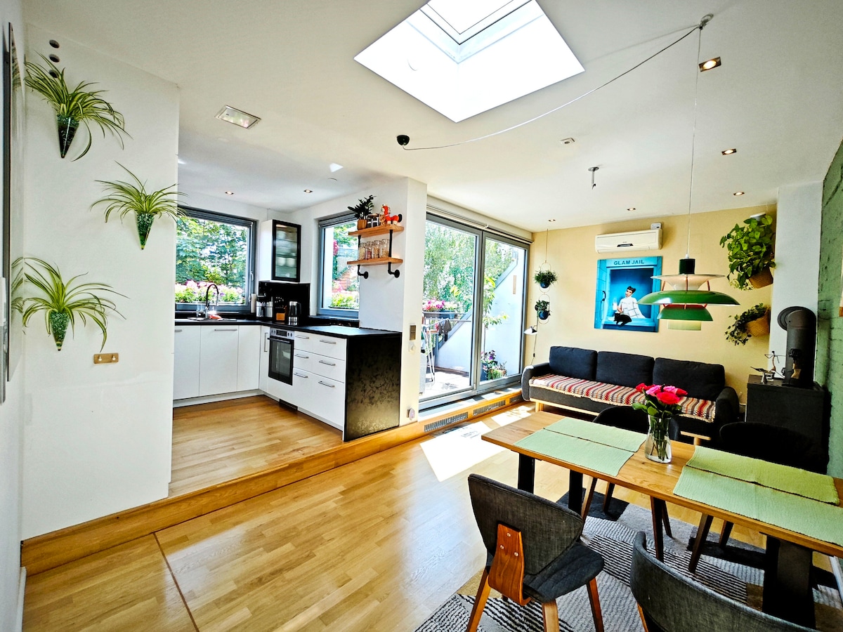 An open living area features a bright, airy kitchen with modern appliances and a spacious dining table. A comfortable sofa faces a wall-mounted television. Large glass doors allow natural light to flood the space, leading to a terrace with greenery visible outside.