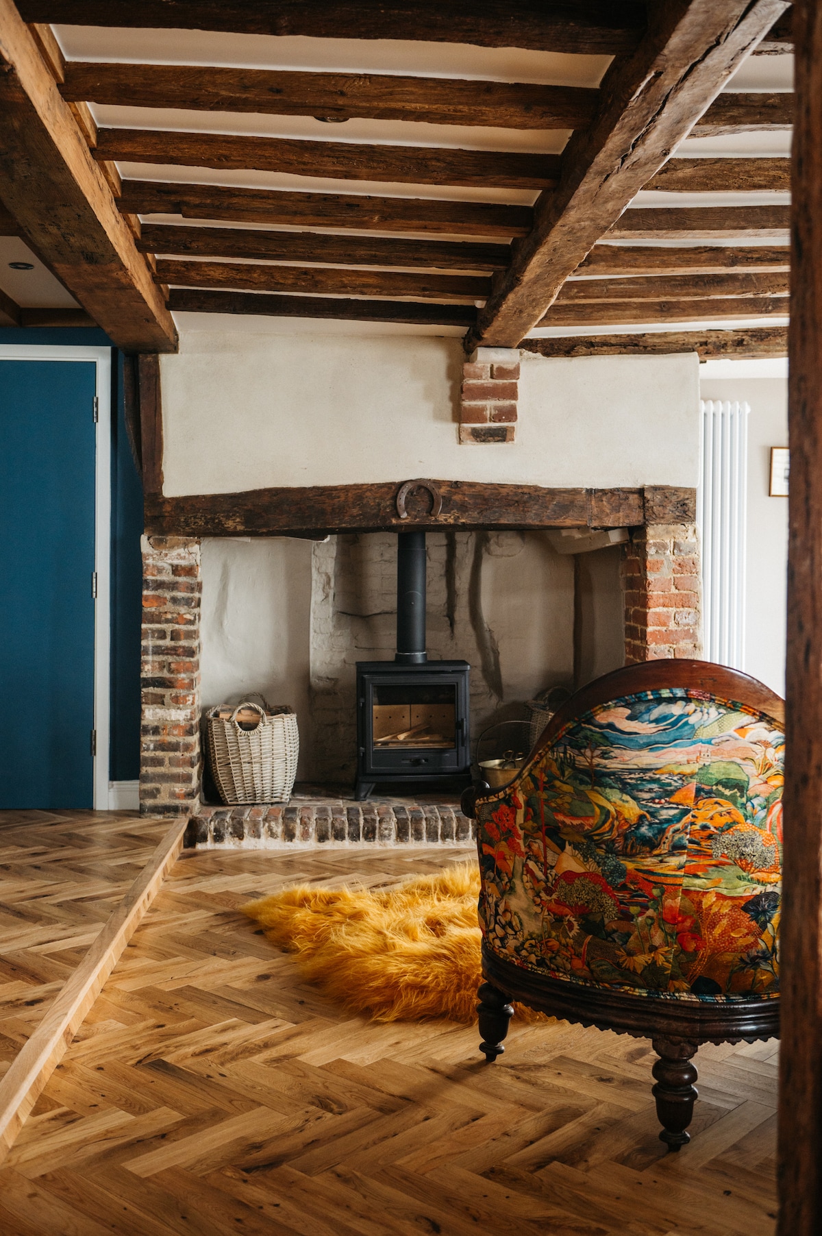 The image features an inviting lounge area with exposed wooden beams and a rustic brick fireplace housing a modern wood-burning stove. A decorative chair with a floral upholstery design is positioned nearby, complemented by a vibrant yellow rug that adds a touch of warmth.