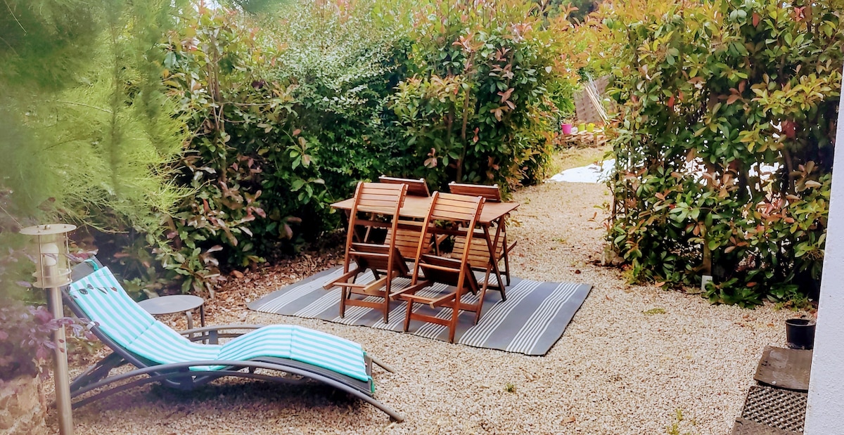 An outdoor space features a gravel surface surrounded by lush greenery. A striped lounge chair is positioned next to a small round table. A set of four folding wooden chairs is arranged around a rectangular mat situated at the center. The atmosphere is calm and private.