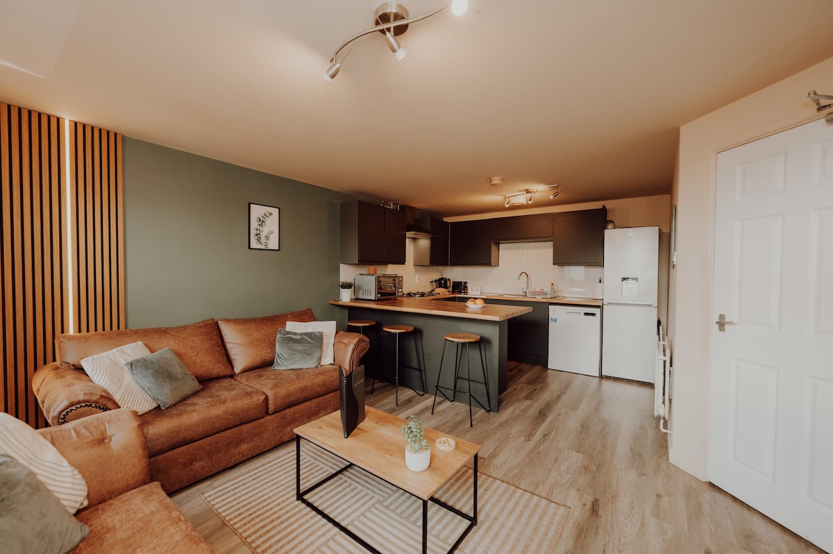 A modern living area features a cozy brown sofa with soft cushions, positioned beside a light wooden coffee table. A fully equipped kitchen is visible in the background, complemented by sleek countertops and contemporary cabinetry. Natural light enters through the window, enhancing the warm tones of the decor.