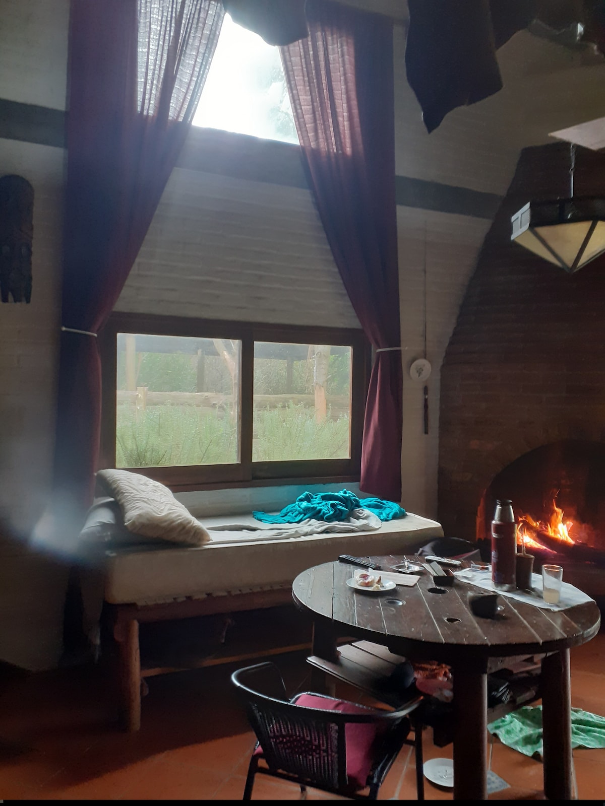 A cozy corner features a rustic wooden table with partially finished drinks and a plate. A comfortable daybed is positioned by a large window, framed with burgundy curtains. A warm fire burns in a brick fireplace, creating an inviting atmosphere.