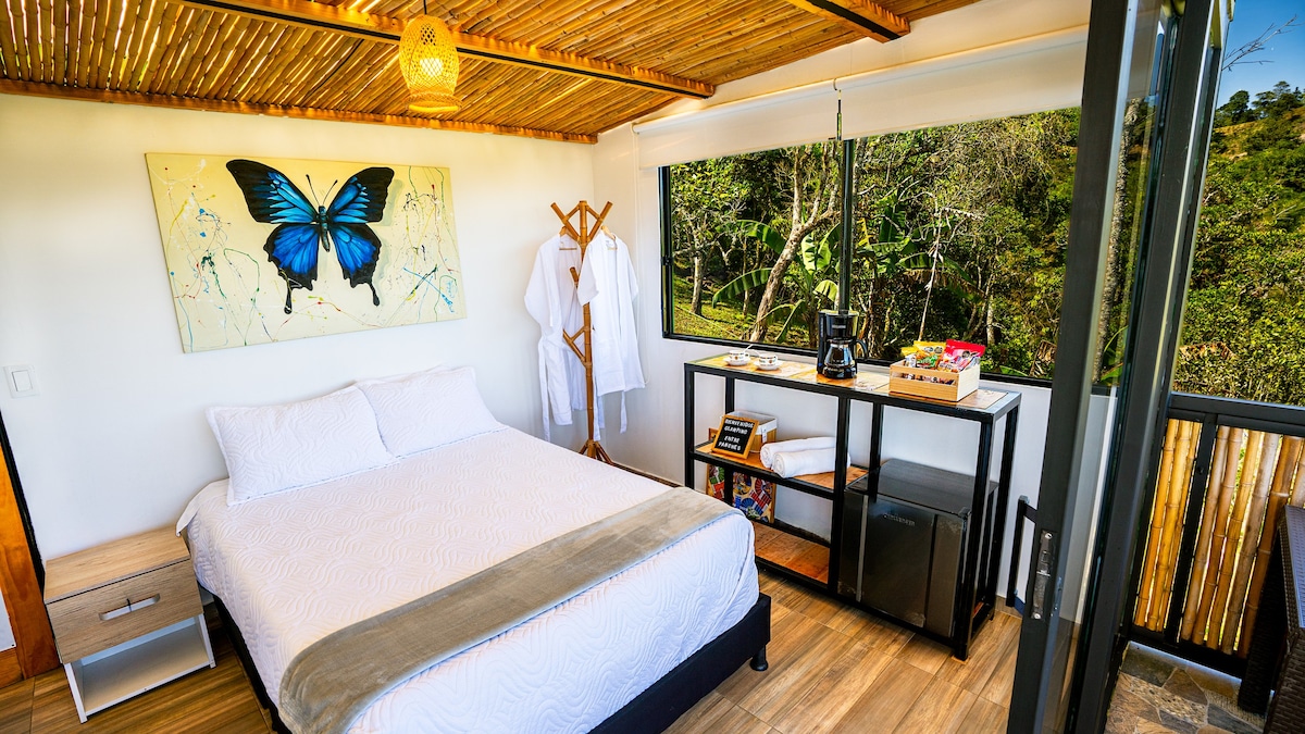 A bright bedroom features a spacious bed with a light-colored bedspread and a decorative butterfly painting above it. Large windows overlook greenery, allowing natural light to fill the room. A small table displays refreshments, while cozy robes hang nearby.