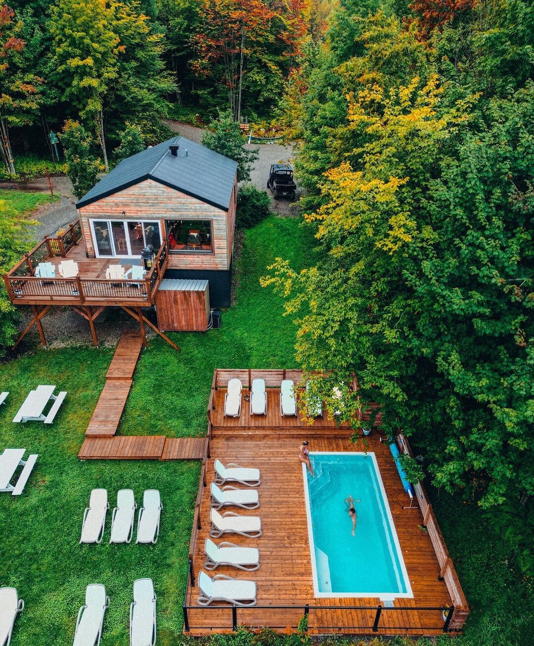 An aerial view of a heated saltwater pool surrounded by wooden decking and lounge chairs, nestled among lush greenery. A spacious cabin with a balcony overlooks the pool, providing a serene setting amidst the vibrant trees.