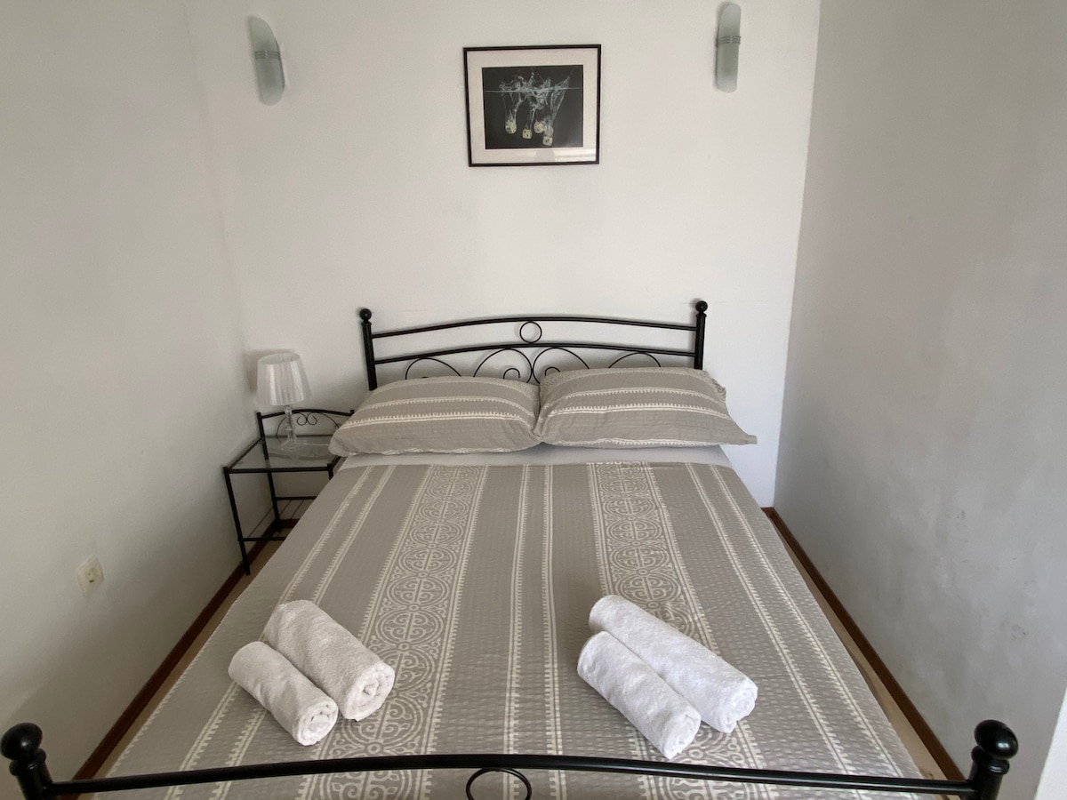 A sleeping area features a metal bed framed with a decorative headboard, dressed in striped bedding. Fluffy towels are neatly arranged on the bed. A small bedside table holds a lamp, and a framed artwork adorns the wall above the bed.