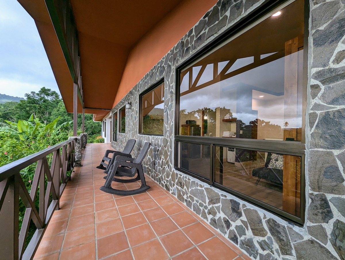 A wide balcony featuring rock-faced walls is adorned with dark wooden rocking chairs. Large windows allow for expansive views of the surrounding lush greenery and hills, creating a relaxing outdoor space.