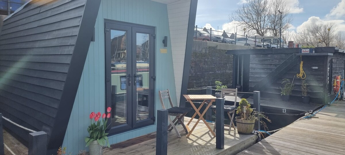 The exterior of the floating glamping pod is showcased, featuring a light blue wall with large glass doors. A wooden deck is visible, furnished with a small table and chairs, complemented by potted plants. The marina and neighboring boats can be seen in the background.