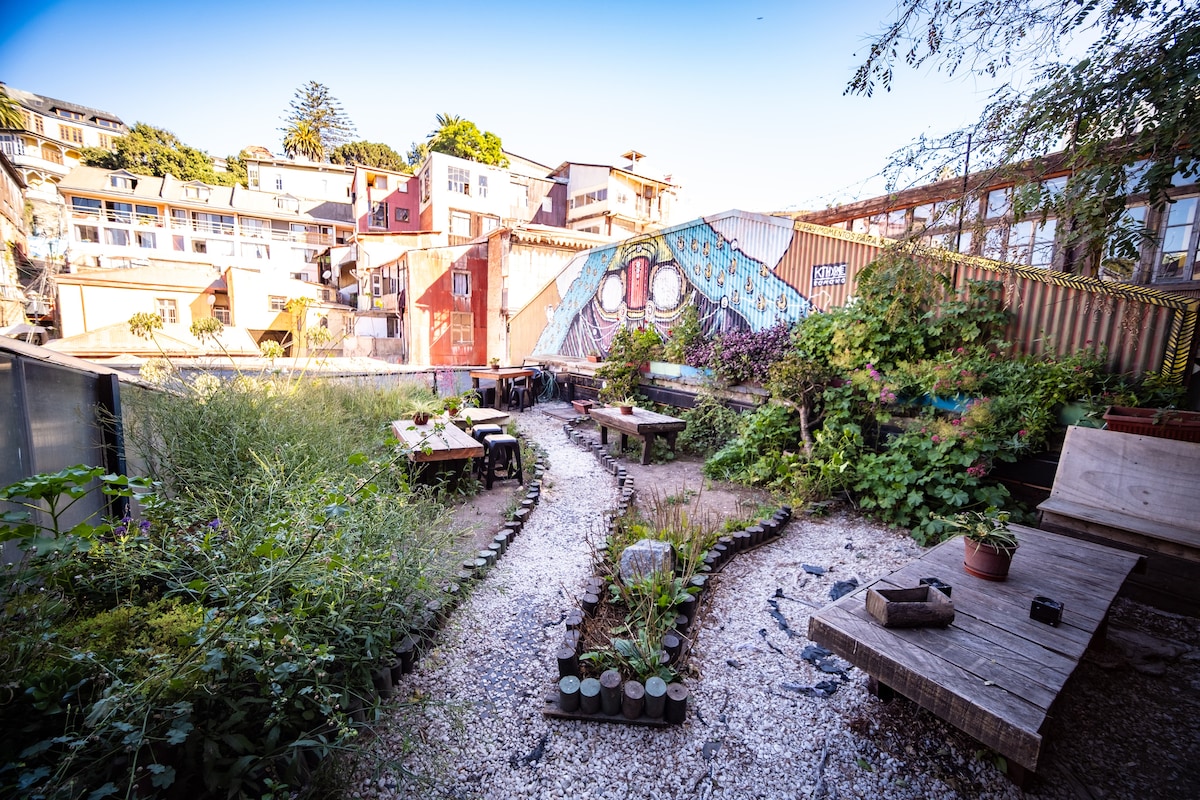 A community garden space is displayed, featuring a winding path bordered by greenery and decorative stones. Several wooden tables are arranged throughout, inviting gathering. Colorful murals adorn nearby walls, contributing to a vibrant atmosphere. The area offers a peaceful retreat among the city landscape.