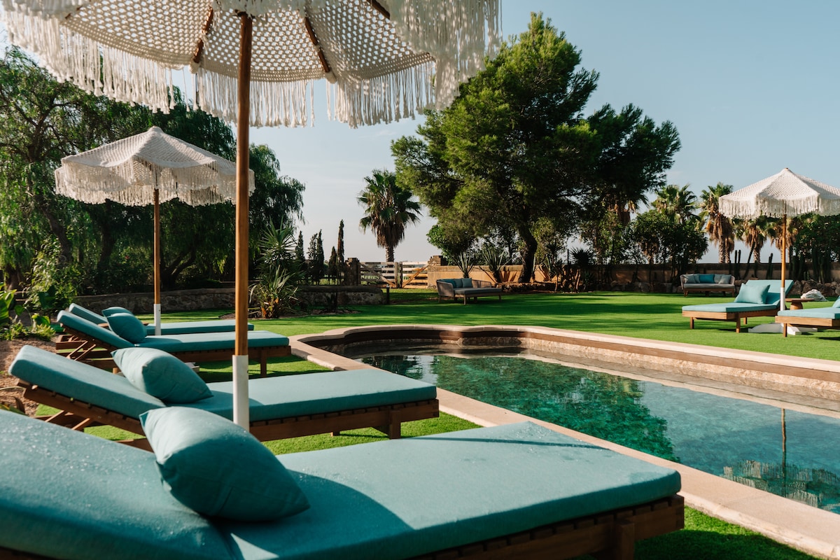 A serene outdoor area features a refreshing pool surrounded by lounge chairs with soft cushions. Umbrellas provide shade, while lush greenery and palm trees enhance the peaceful environment. The space invites relaxation under clear skies, perfect for enjoying Mallorca's warm climate.