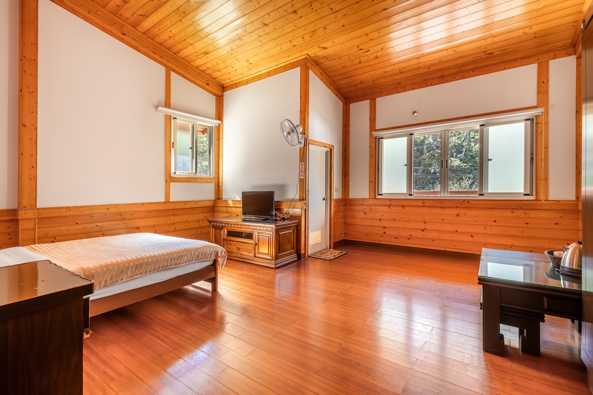 A spacious room with wooden walls and ceiling features a simple bed, a TV on an elegant stand, and a small table. Two large windows allow natural light to brighten the area, while a door provides access to an outdoor view.