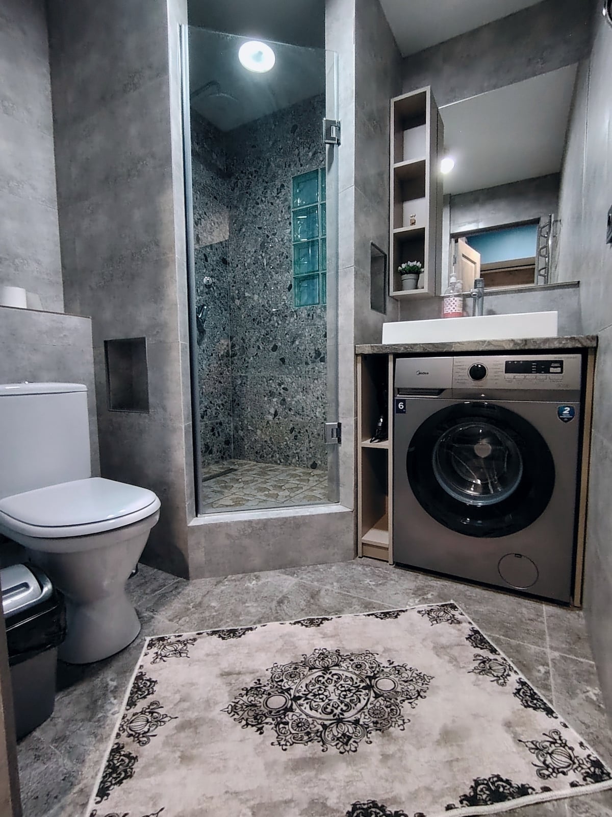 A modern bathroom features a sleek glass shower with textured tile walls. A compact washing machine is integrated into cabinetry alongside a stylish sink and a color-coordinated rug lies on the floor, adding a decorative touch to the space.