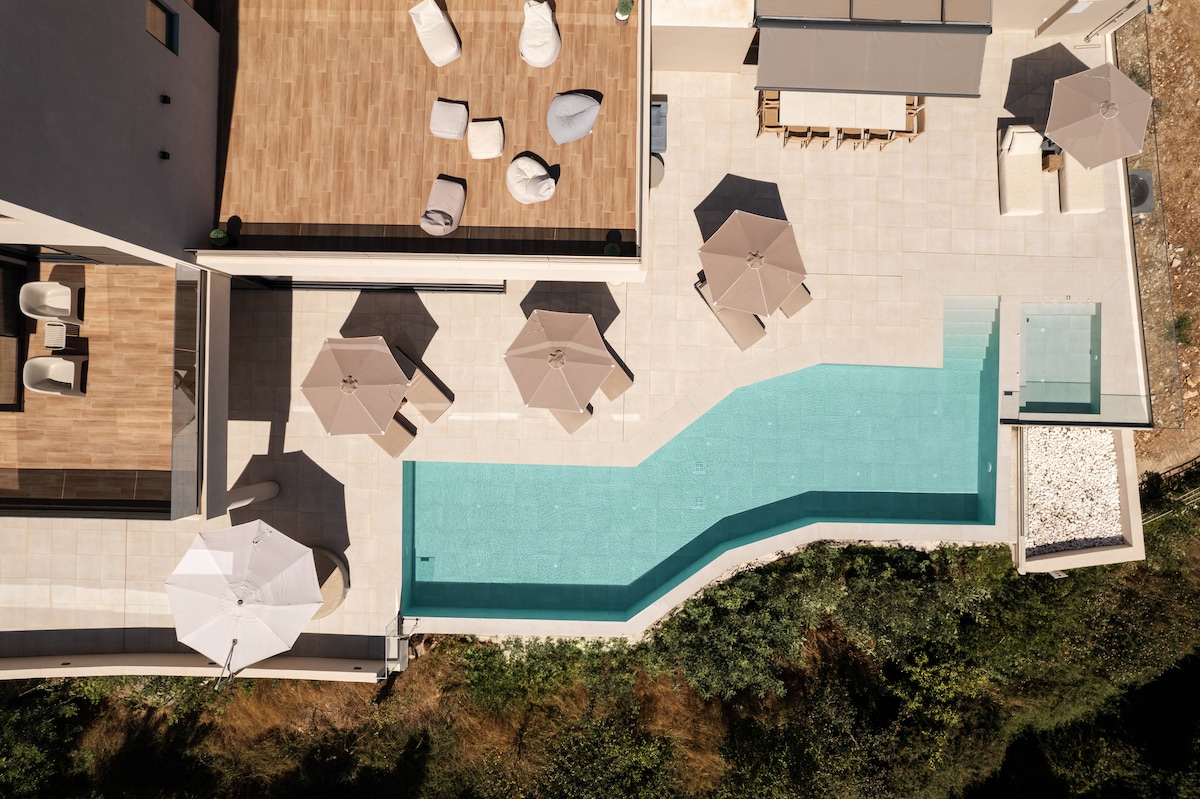 An aerial view showcases a uniquely shaped swimming pool surrounded by sunbeds and umbrellas. The outdoor area features a spacious terrace with cushioned loungers and seating arrangements under shaded umbrellas, inviting relaxation and enjoyment in a tranquil atmosphere.