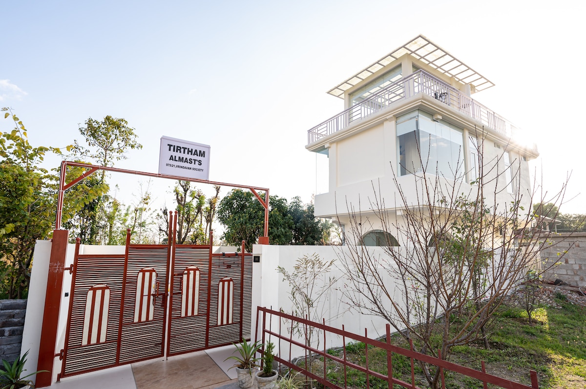 The entrance to Tirtham features a welcoming gate with the name 'Tirtham' prominently displayed. A modern, three-story villa is visible beyond the gate, surrounded by well-maintained greenery. Natural light highlights the villa's architectural details against a clear sky.