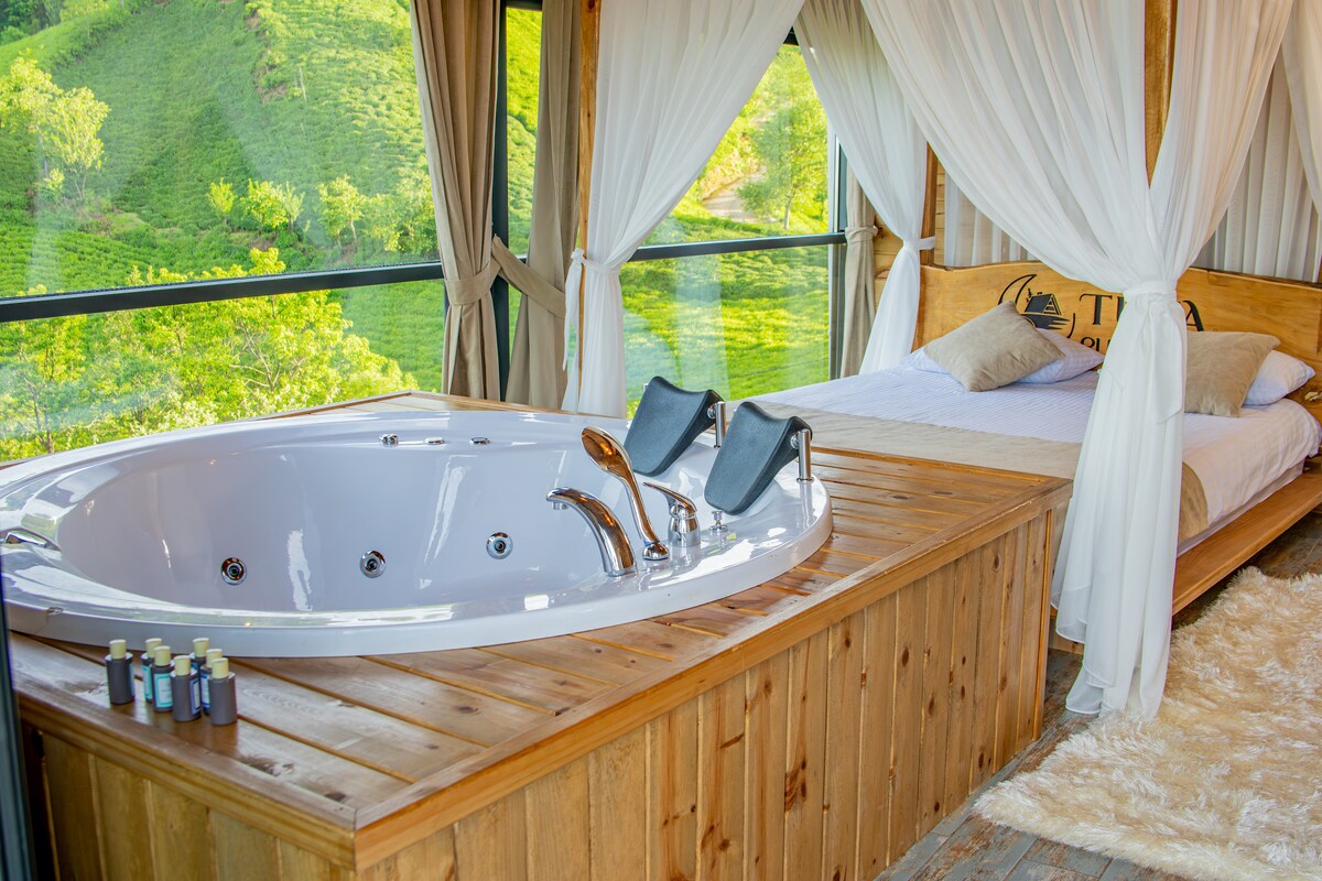 A wooden platform features a spacious jacuzzi, flanked by two reclining chairs. Soft drapes provide a sense of privacy, while a cozy sleeping area is positioned nearby with light-colored bedding. Glimpses of lush green hills are visible through large glass windows.