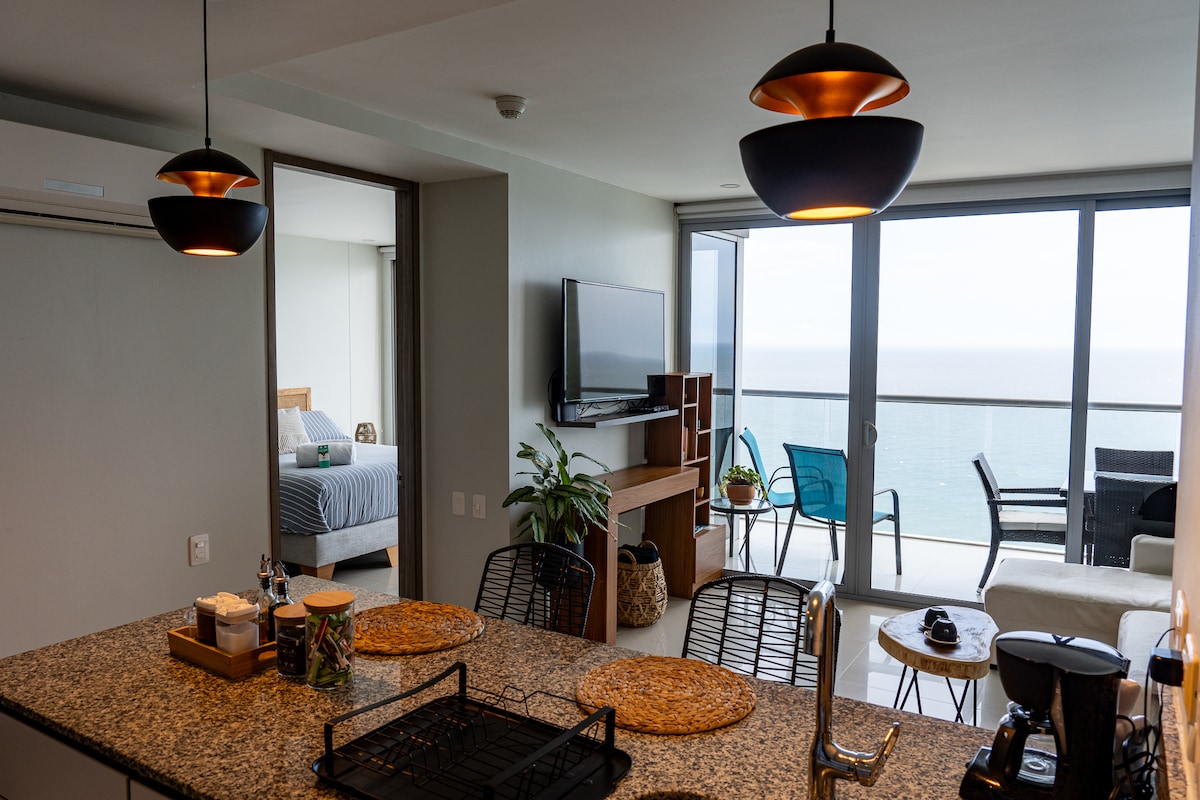 A modern open-concept living area is shown, featuring a well-equipped kitchen with granite countertops and seating. The space is enhanced by large windows, offering views of the sea. A glimpse of the bedroom and a cozy lounge area with blue chairs can be seen in the background.