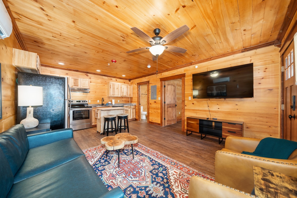 A living area with warm wooden walls and ceiling is featured. A blue couch is positioned alongside a rustic coffee table. A flat-screen TV is mounted on one wall, complemented by a mounted ceiling fan. The kitchen area is visible in the back, showing wooden cabinetry and appliances.