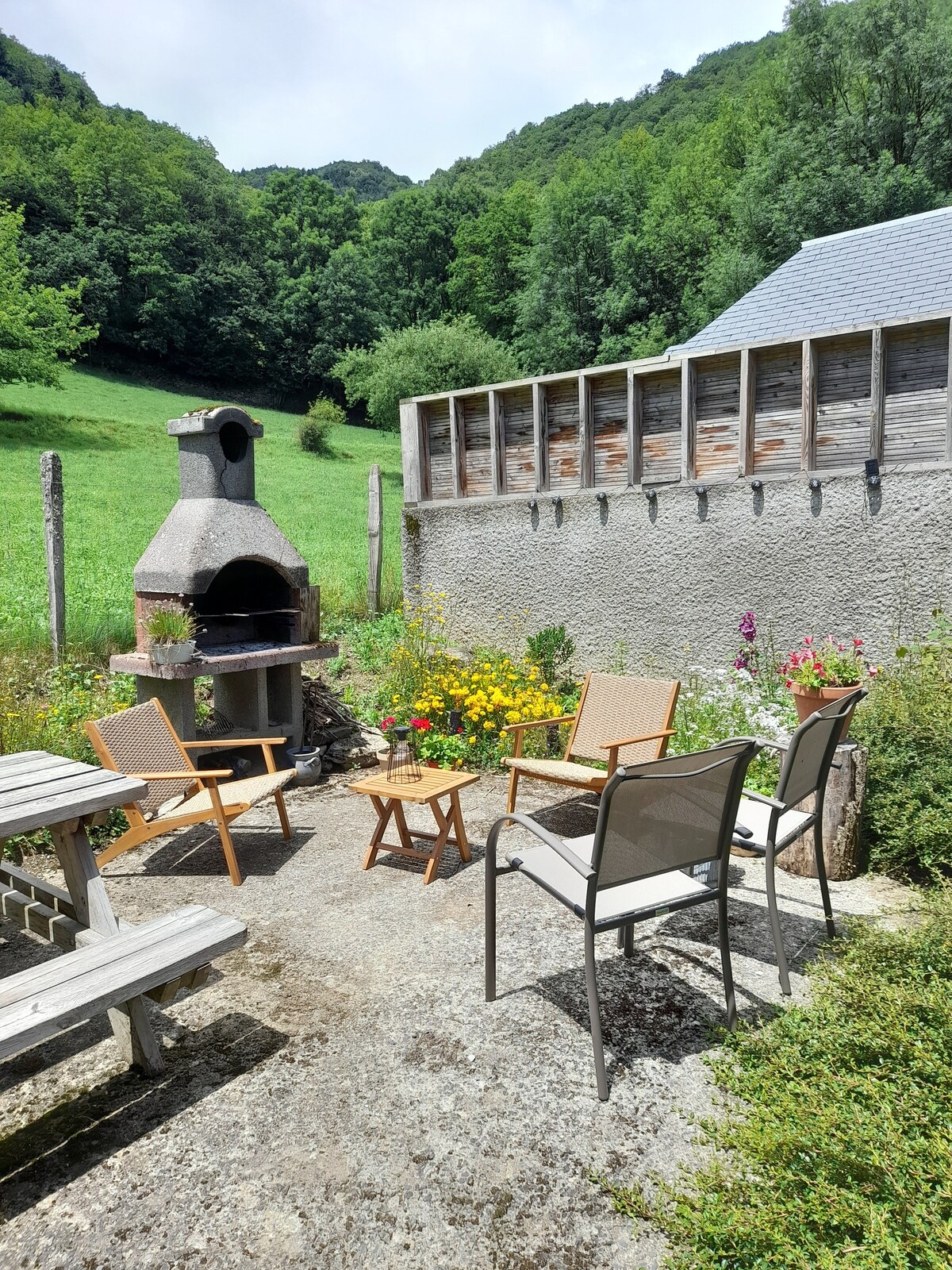 An outdoor patio area features a stone barbecue and a small wooden table surrounded by four chairs. Vibrant flowers and greenery provide a natural backdrop, while a wooden structure is seen along the back, enhancing the tranquil setting.