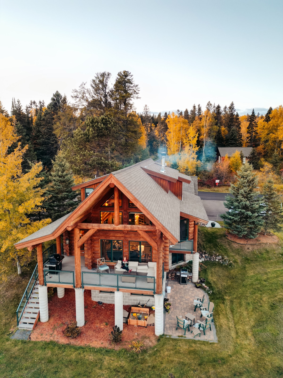 The log home is set amidst lush greenery and autumn foliage. The spacious deck features comfortable seating and overlooks the landscaped yard. A sloped roof and large windows enhance the rustic charm, while the surrounding trees add natural privacy.