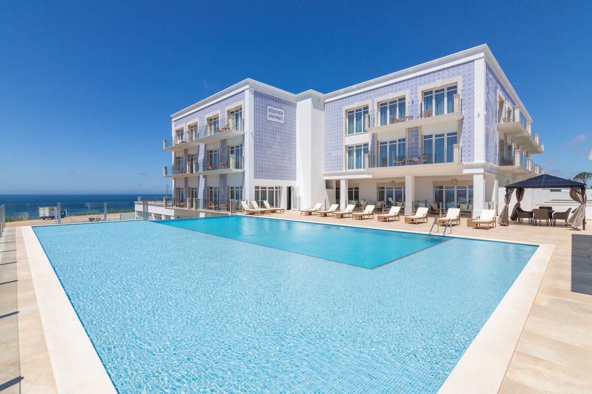 A modern resort building features a spacious outdoor pool, surrounded by lounge chairs and offering sweeping views of the ocean. The architectural design includes large windows and balconies, allowing natural light to flood the space, and emphasizes the inviting atmosphere of the resort.