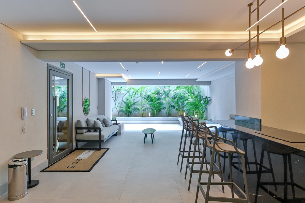 A modern common area features a sleek bar counter with high stools and a minimalistic table. Natural light filters in through large windows, illuminating the area and highlighting the greenery of indoor plants in a calming atmosphere.
