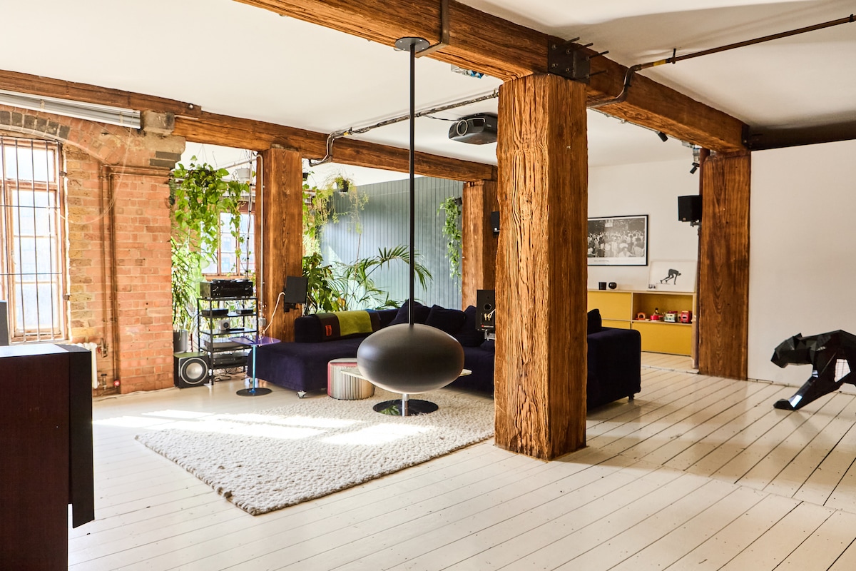 A spacious living area features exposed wooden beams and light-coloured wooden flooring. A dark purple sofa is arranged around a soft area rug, complemented by greenery positioned along the walls. A unique hanging light fixture adds visual interest, while entertainment equipment is discreetly included.