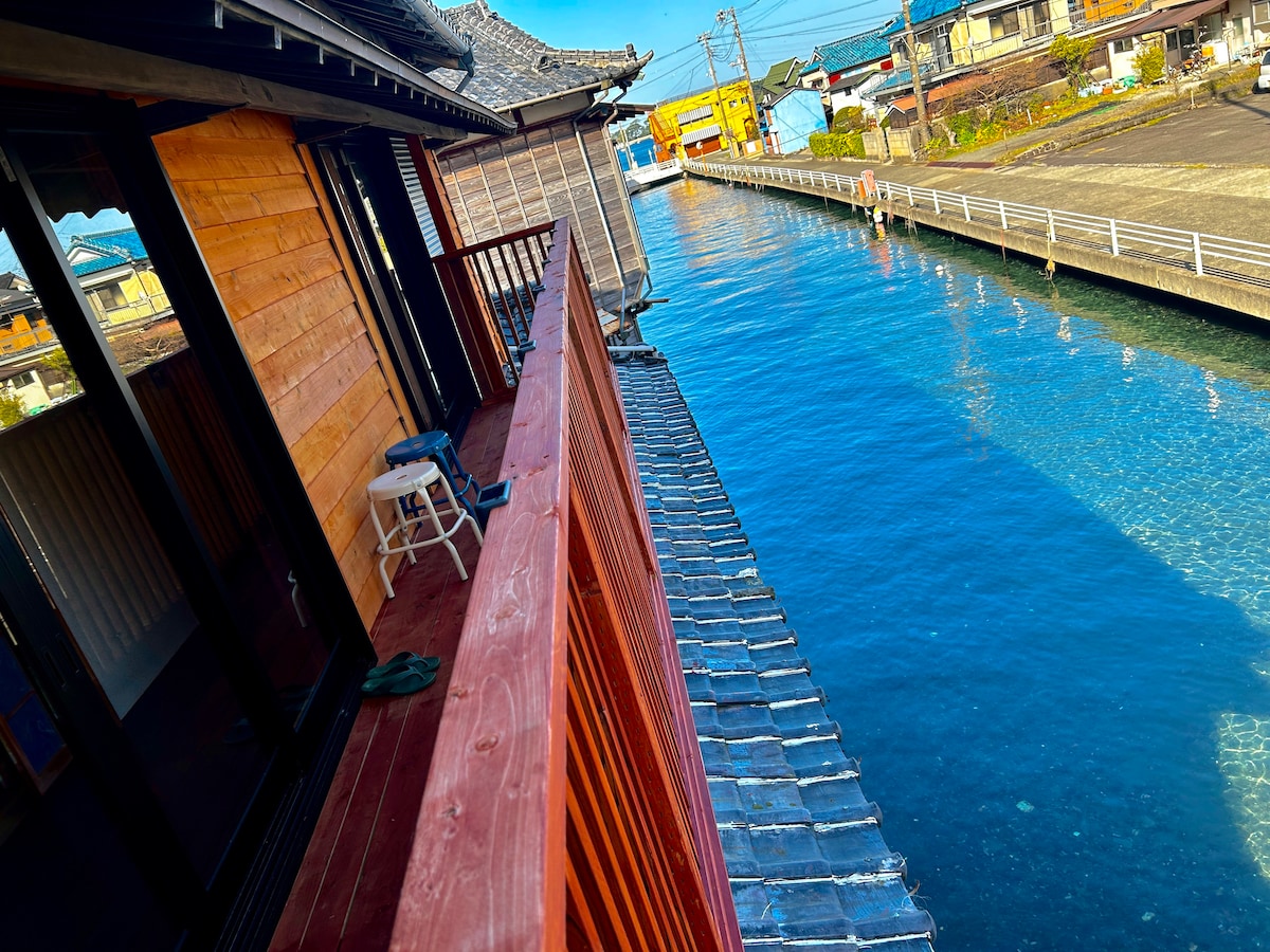A spacious balcony offers a view of the clear blue river below. The wooden railing aligns with the natural wooden siding of the house. Nearby, the shoreline is visible, showcasing a pathway that runs alongside the water and hints at the tranquility of the surroundings.